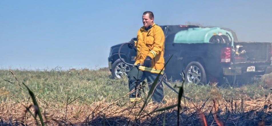 Firefighter using a hose to extinguish a grass fire, with a pickup truck carrying a water tank in the background. Firefighter using a hose to extinguish a grass fire, with a pickup truck carrying a water tank in the background.