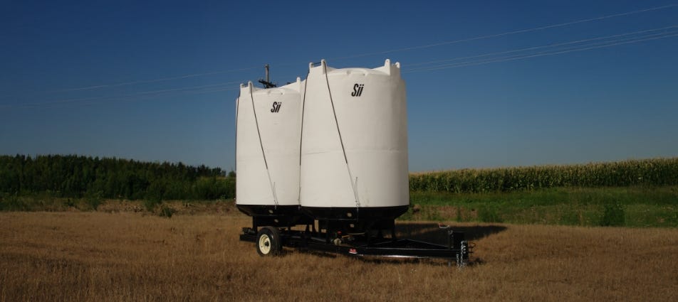 Two white double-wall storage tanks mounted on a trailer in a farm field near a corn crop. Two white double-wall storage tanks mounted on a trailer in a farm field near a corn crop.