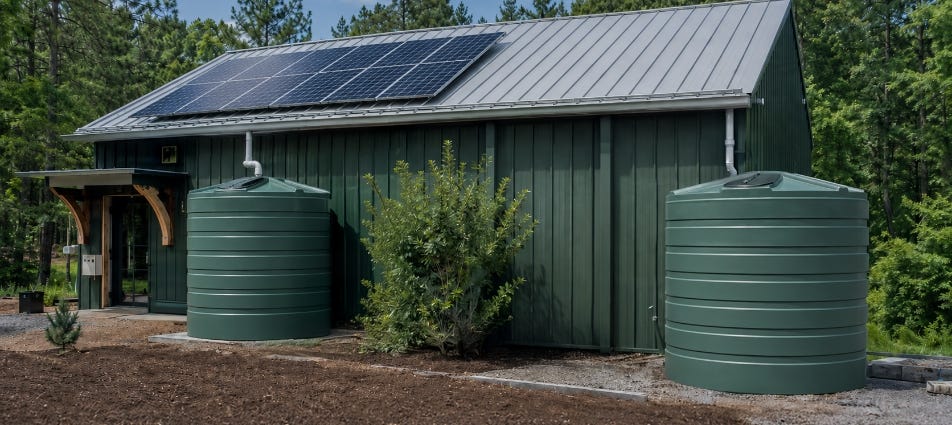 Two green rainwater storage tanks connected to gutters beside a metal-roof building with solar panels in a wooded setting. Two green rainwater storage tanks connected to gutters beside a metal-roof building with solar panels in a wooded setting.