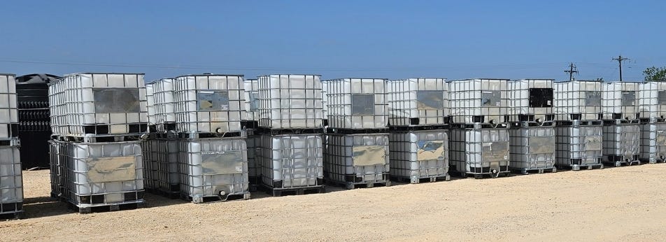 Dozens of white intermediate bulk containers (IBC totes) with metal cages, stacked two high in long rows outdoors on a light gravel yard under a clear blue sky. Dozens of white intermediate bulk containers (IBC totes) with metal cages, stacked two high in long rows outdoors on a light gravel yard under a clear blue sky.