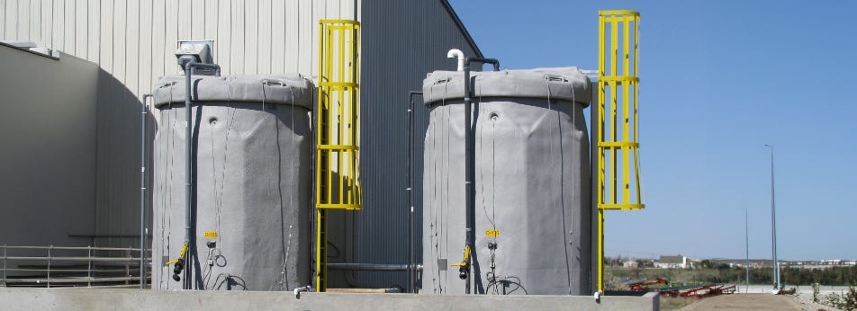 Two gray industrial storage tanks with piping and yellow access ladders installed beside a metal building. Two gray industrial storage tanks with piping and yellow access ladders installed beside a metal building.