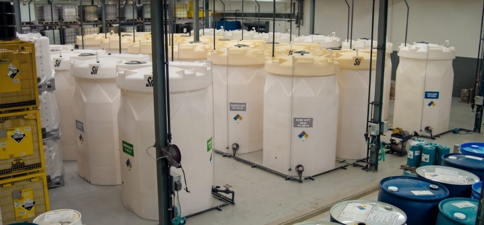 Indoor chemical storage area with multiple white industrial tanks connected by piping and surrounded by labeled chemical containers. Indoor chemical storage area with multiple white industrial tanks connected by piping and surrounded by labeled chemical containers.