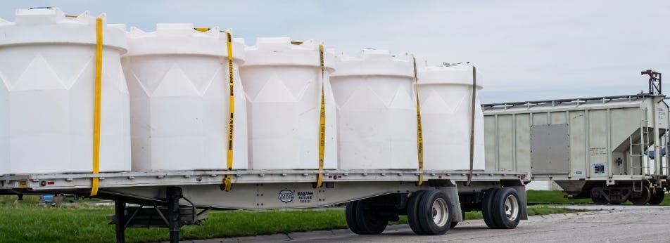 White industrial chemical storage tanks secured on a flatbed trailer for transport near an industrial rail facility. White industrial chemical storage tanks secured on a flatbed trailer for transport near an industrial rail facility.