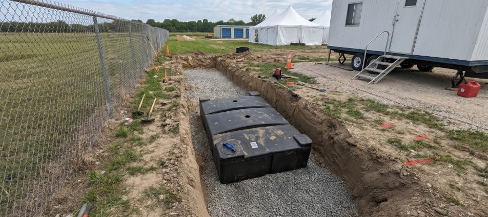 Black rectangular septic tank installed in a gravel-lined trench at a temporary jobsite with a portable office trailer nearby. Black rectangular septic tank installed in a gravel-lined trench at a temporary jobsite with a portable office trailer nearby.