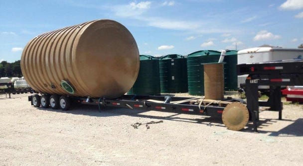 Large fiberglass storage tank secured on a flatbed trailer for nationwide delivery, with additional storage tanks visible in the background. Large fiberglass storage tank secured on a flatbed trailer for nationwide delivery, with additional storage tanks visible in the background.