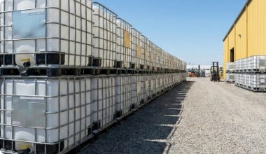 Rows of stacked caged IBC totes stored in an outdoor bulk order yard beside an industrial warehouse. Rows of stacked caged IBC totes stored in an outdoor bulk order yard beside an industrial warehouse.