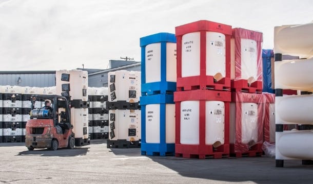 Forklift moving stacked chemical IBC totes in an industrial storage yard with red, blue, and white framed containers. Forklift moving stacked chemical IBC totes in an industrial storage yard with red, blue, and white framed containers.