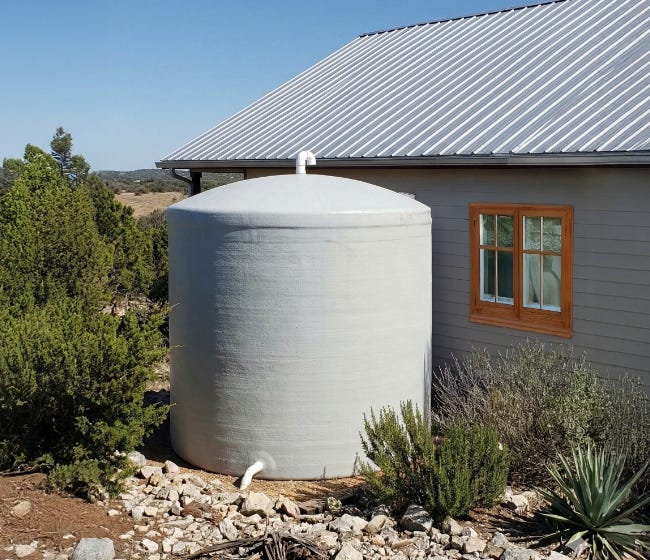 Gray fiberglass rainwater storage tank installed beside a house with a metal roof in a landscaped residential setting. Gray fiberglass rainwater storage tank installed beside a house with a metal roof in a landscaped residential setting.