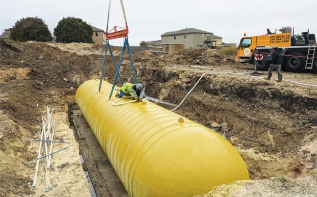 Large yellow underground storage tank being lowered into an excavated trench by a crane at a construction site. Large yellow underground storage tank being lowered into an excavated trench by a crane at a construction site.