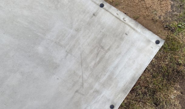 Close-up top view of an Instaslab foundation mat corner secured with black pegs on a prepared outdoor surface. Close-up top view of an Instaslab foundation mat corner secured with black pegs on a prepared outdoor surface.