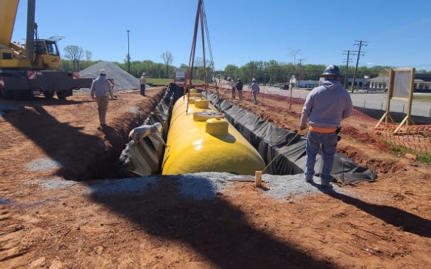 Yellow underground fiberglass storage tank being lowered into a lined excavation trench with workers and crane equipment on site. Yellow underground fiberglass storage tank being lowered into a lined excavation trench with workers and crane equipment on site.