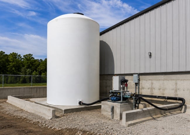White vertical brine storage tank connected to pumping and transfer equipment beside an industrial building. White vertical brine storage tank connected to pumping and transfer equipment beside an industrial building.