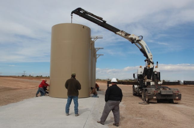 Vertical fiberglass fire suppression water tank being installed on a concrete pad with crane equipment and workers at a rural jobsite. Vertical fiberglass fire suppression water tank being installed on a concrete pad with crane equipment and workers at a rural jobsite.