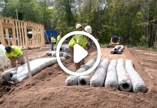Workers installing EZflow drainfield bundles at a residential septic leach field site with construction framing and equipment in the background. Workers installing EZflow drainfield bundles at a residential septic leach field site with construction framing and equipment in the background.