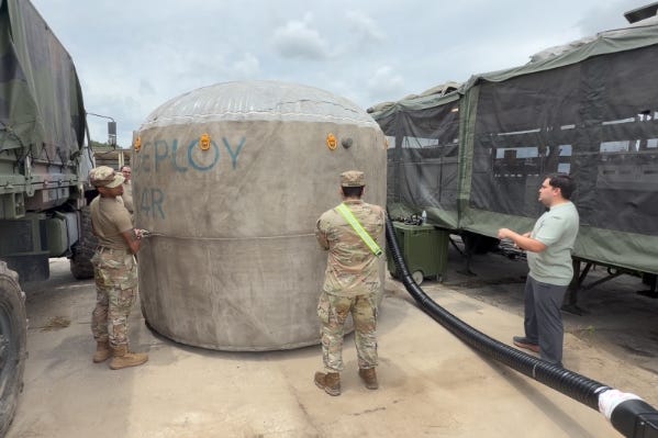 Military personnel and a technician inspecting an inflated concrete Omni Tank connected to a large hose between transport trucks. Military personnel and a technician inspecting an inflated concrete Omni Tank connected to a large hose between transport trucks.