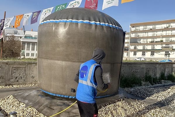 Worker applying water to a deployed concrete Omni Tank on an Instaslab foundation mat in an urban outdoor setting. Worker applying water to a deployed concrete Omni Tank on an Instaslab foundation mat in an urban outdoor setting.