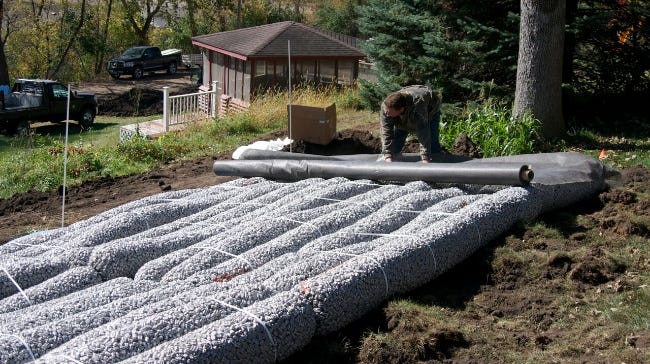 Worker installing EZflow drainfield bundles on a sloped residential site with a perforated pipe being placed across the system. Worker installing EZflow drainfield bundles on a sloped residential site with a perforated pipe being placed across the system.