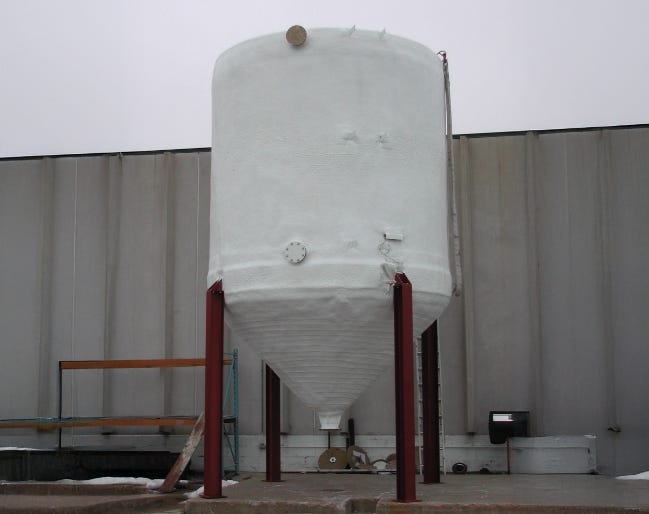 White industrial FRP cone-bottom storage tank mounted on steel support legs beside a facility wall. White industrial FRP cone-bottom storage tank mounted on steel support legs beside a facility wall.