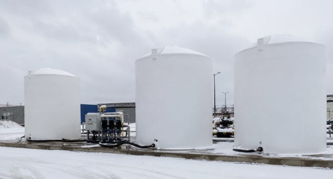 White vertical brine storage tanks installed outdoors at a snowy industrial facility with connected pumping equipment. White vertical brine storage tanks installed outdoors at a snowy industrial facility with connected pumping equipment.
