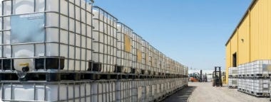 Rows of stacked caged IBC totes stored in an outdoor bulk order yard beside an industrial warehouse. Rows of stacked caged IBC totes stored in an outdoor bulk order yard beside an industrial warehouse.