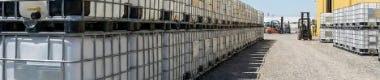 Rows of stacked caged IBC totes stored in an outdoor bulk order yard beside an industrial warehouse. Rows of stacked caged IBC totes stored in an outdoor bulk order yard beside an industrial warehouse.