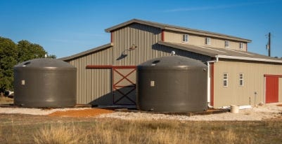 Two large fiberglass rainwater storage tanks installed beside a metal barn for agricultural water collection and storage. Two large fiberglass rainwater storage tanks installed beside a metal barn for agricultural water collection and storage.