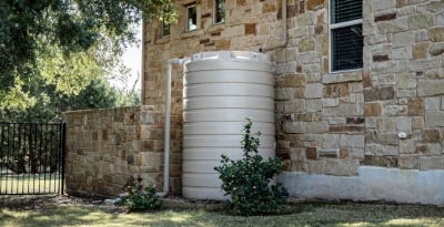Beige poly rainwater tank installed beside a stone house for residential water collection and storage. Beige poly rainwater tank installed beside a stone house for residential water collection and storage.