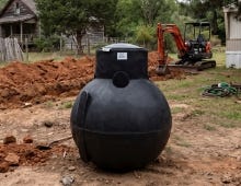 Black septic pump tank placed on a residential jobsite with excavated soil and construction equipment in the background. Black septic pump tank placed on a residential jobsite with excavated soil and construction equipment in the background.