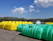 Rows of green and yellow single-compartment septic tanks stored outdoors on a paved lot under a partly cloudy sky. Rows of green and yellow single-compartment septic tanks stored outdoors on a paved lot under a partly cloudy sky.