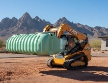 Skid steer loader transporting a large green two-compartment septic tank on a construction site with mountains in the background. Skid steer loader transporting a large green two-compartment septic tank on a construction site with mountains in the background.